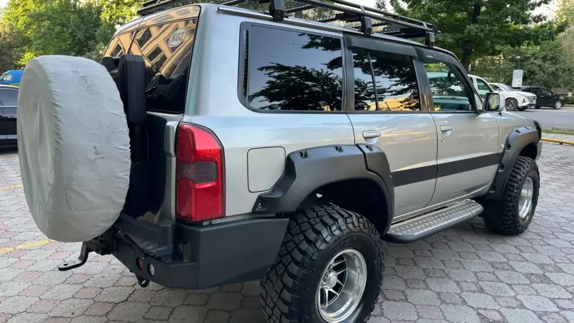 Gray off-road SUV with a roof rack and large mud-terrain tires, parked in a lot; spare tire is covered with a gray protective cover on the rear.