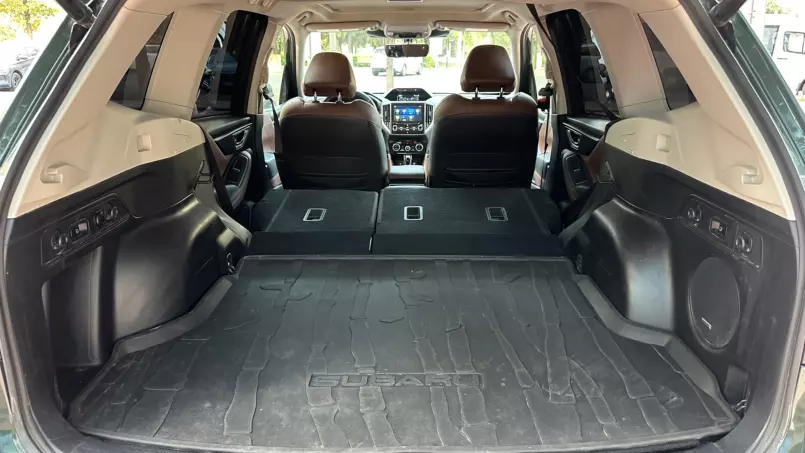 Open hatchback trunk with folded rear seats, creating a large cargo area lined by a black all-weather mat in a Subaru SUV; front seats and center console visible ahead.