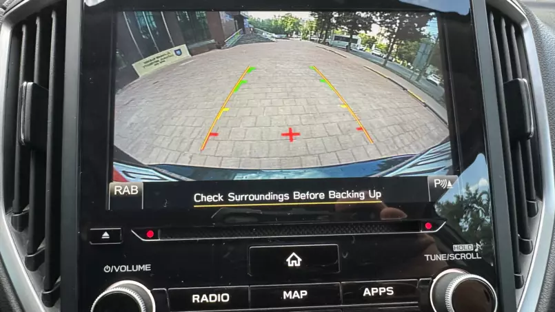 Rearview camera display showing a brick street with trees, guiding lines (green/yellow/red) for backing up; text reads 'Check Surroundings Before Backing Up'.
