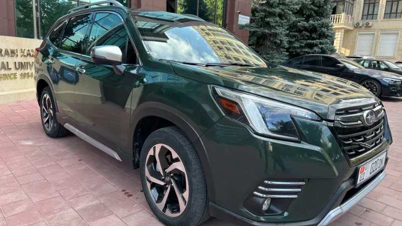 Green SUV parked on a brick sidewalk in front of a glass-front building, with trees reflected in the windows behind it.