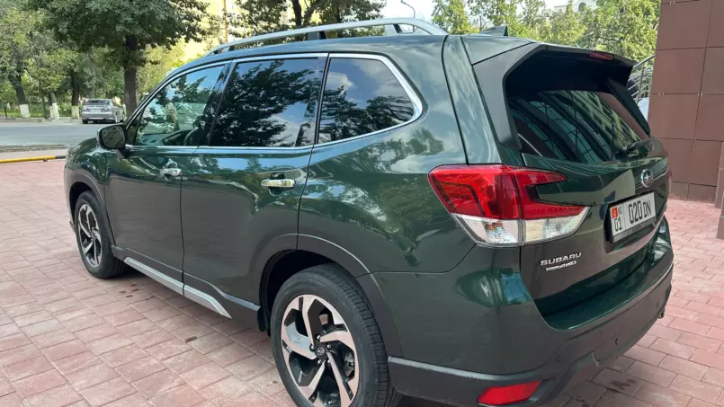 Rear three-quarter view of a dark green Subaru SUV with roof rails parked on a brick surface amid trees.